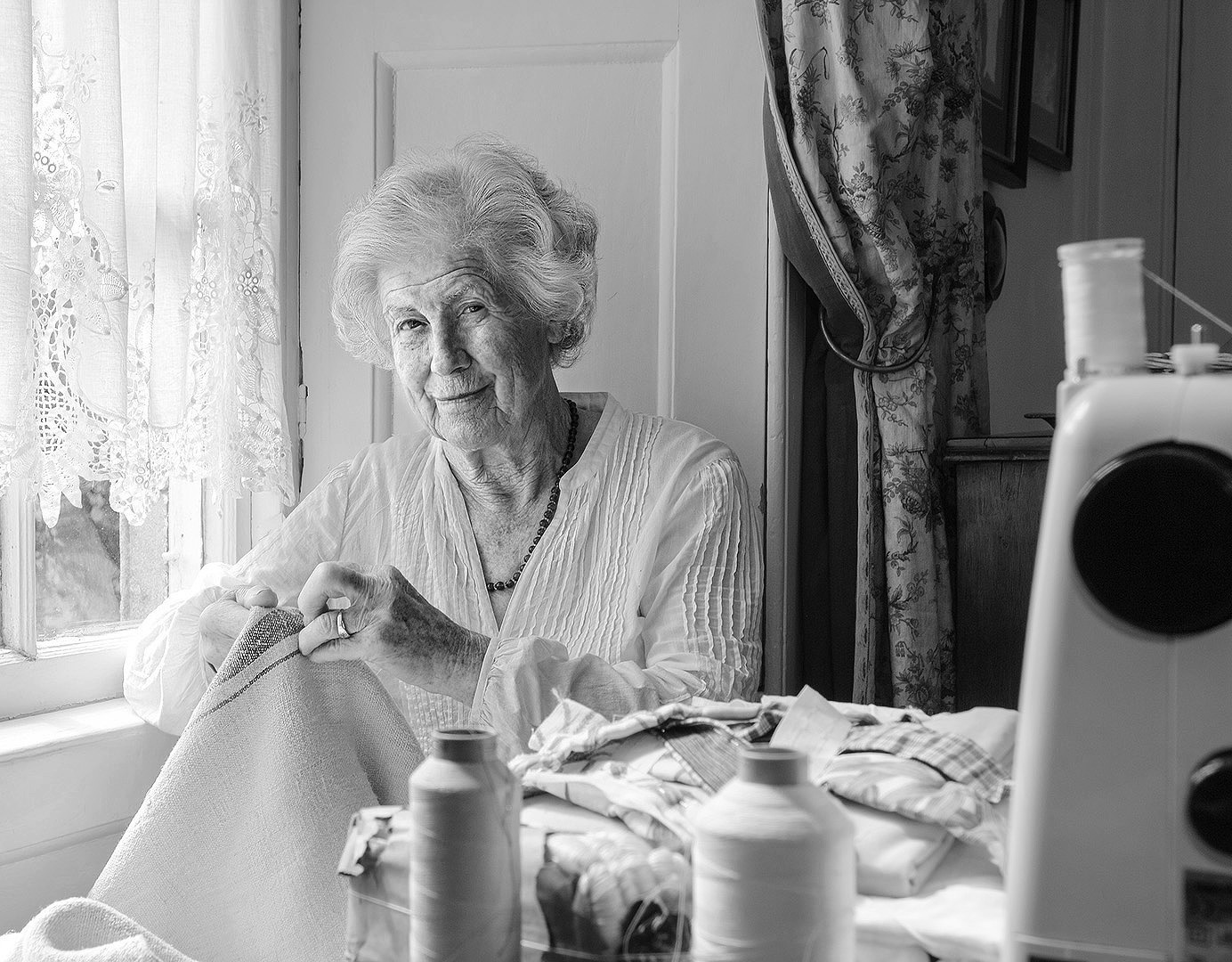B/W photographic portrait of professional clothes maker older woman at window with sewing